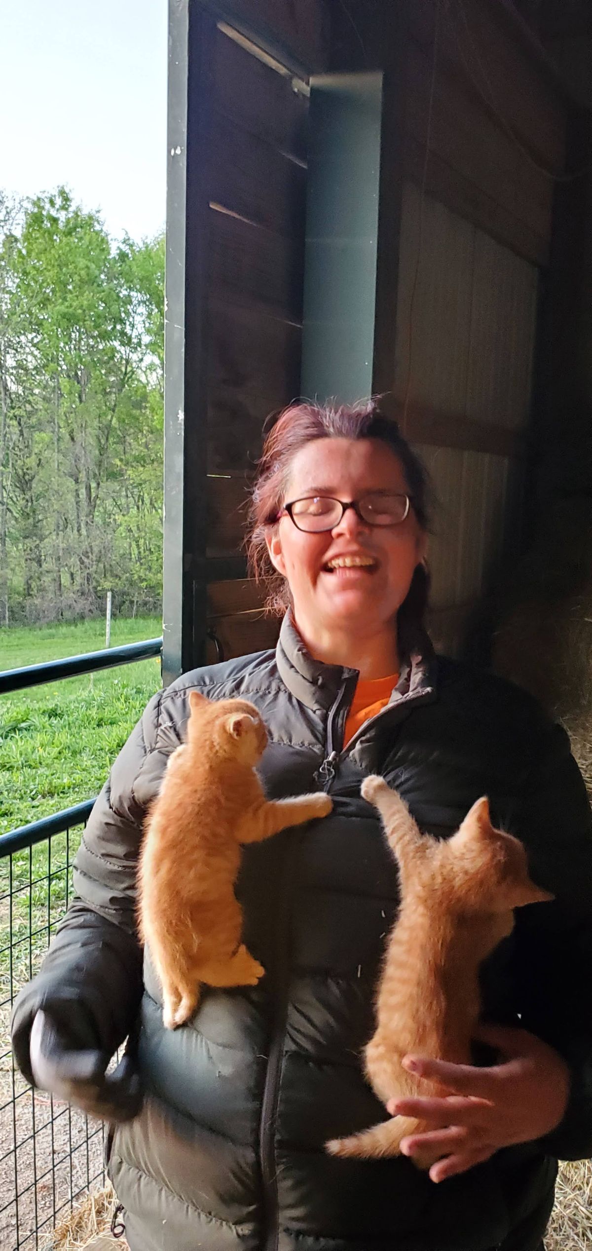 Pure joy radiates from this moment -- Laura beaming with two adorable orange kittens cradled in her arms, the lush green farm stretching out behind her on a beautiful spring day.